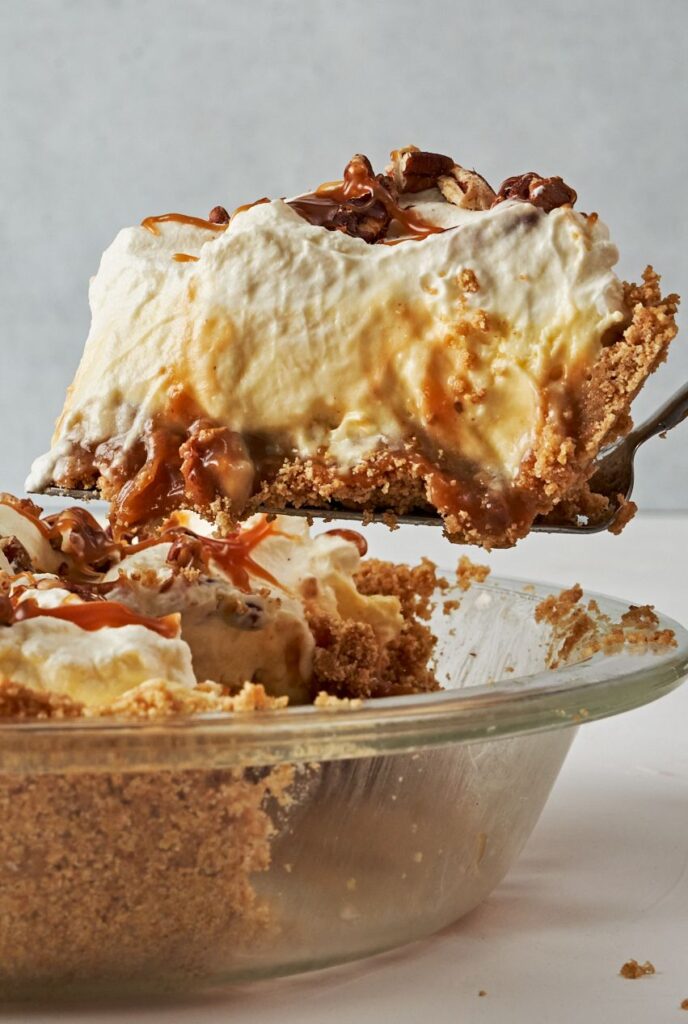 An image of a No-Bake Banana Caramel Pecan Pie placed on a neutral-colored plate. The pie has a prominent, creamy white whipped cream topping, piped to a peak in the center. Beneath the whipped cream, visible layers include golden caramel drizzled over banana slices, which rest on a crumbly graham cracker crust. A few whole pecans are scattered atop the caramel layer. The setting is minimalist, with a soft grey background that accentuates the pie's colors and textures, accompanied by a banana and a small bowl of caramel sauce to the side, suggesting the ingredients used in the pie.